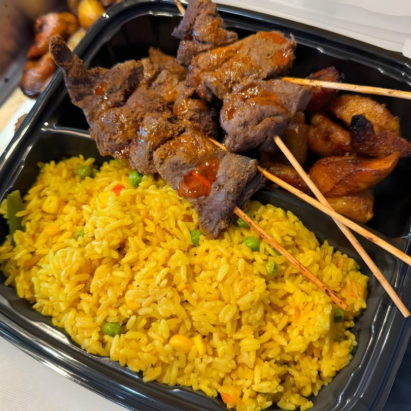 Nigerian meal with yellow rice, beef suya skewers, and fried plantains in a black takeout container.