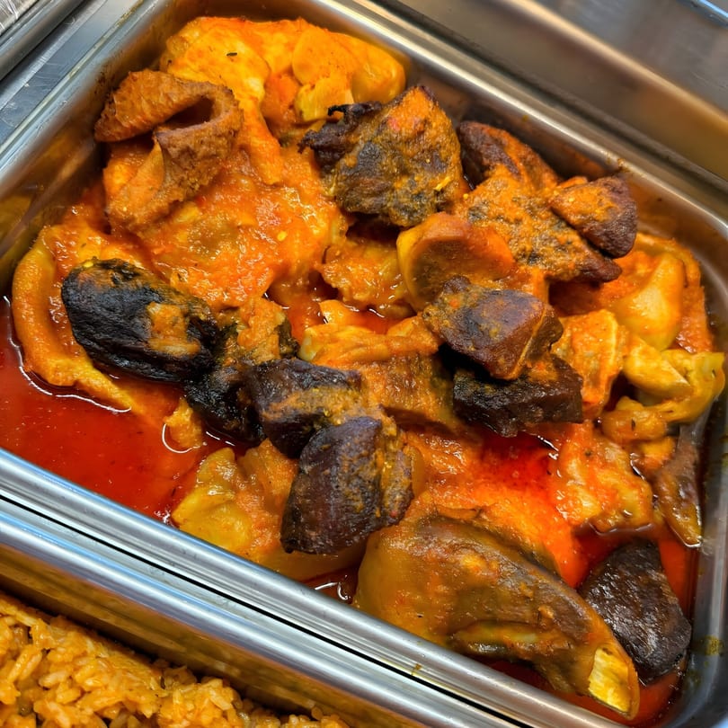 Nigerian stew with assorted meats and vegetables in a metal serving tray, with jollof rice visible below.