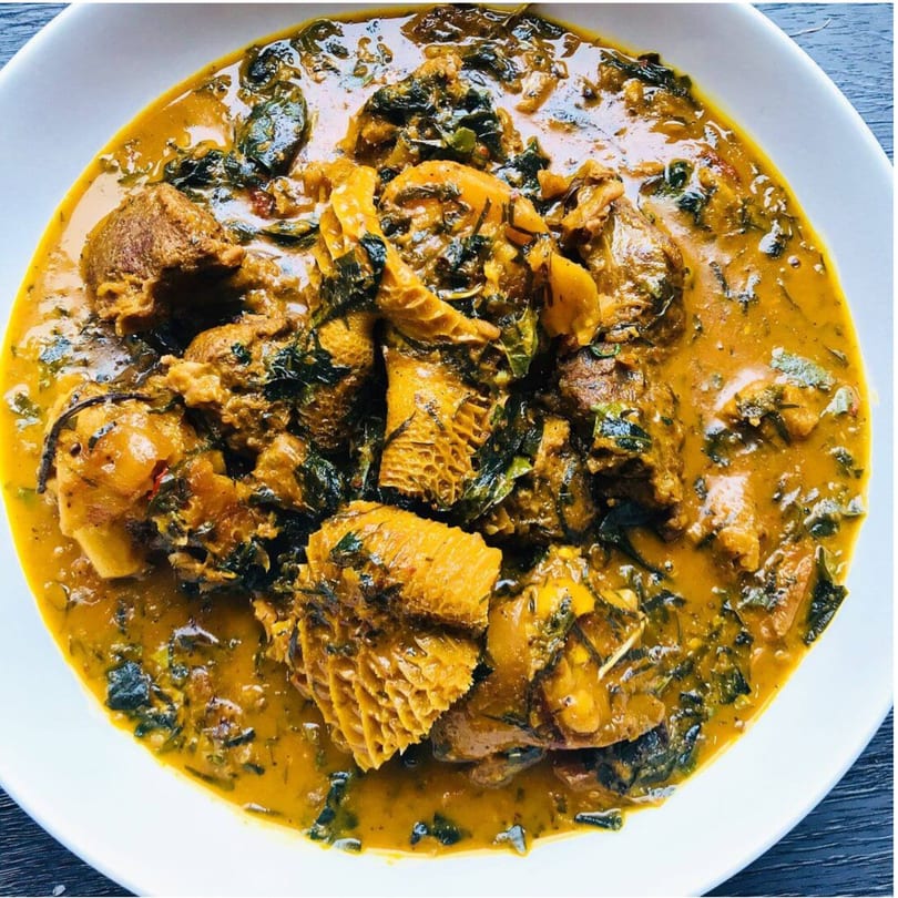A close-up of a Nigerian stew with meat, tripe, and dark green leafy vegetables in a white bowl.