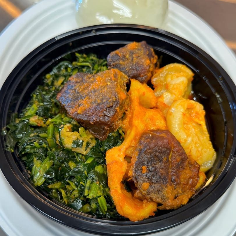 A Nigerian dish with stewed meat, green vegetables, and yellow yam in a black takeout container.