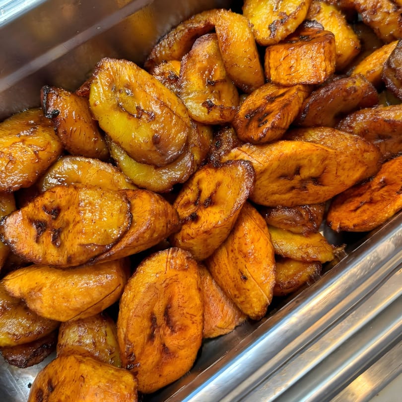 Close-up of golden-brown fried plantains, glistening with oil, in a metal serving tray.